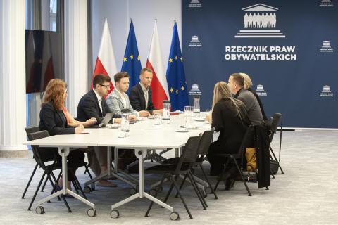 Siedem osób rozmawia przy stole w sali konferencyjnej. W tle flagi Polski i Unii Europejskiej